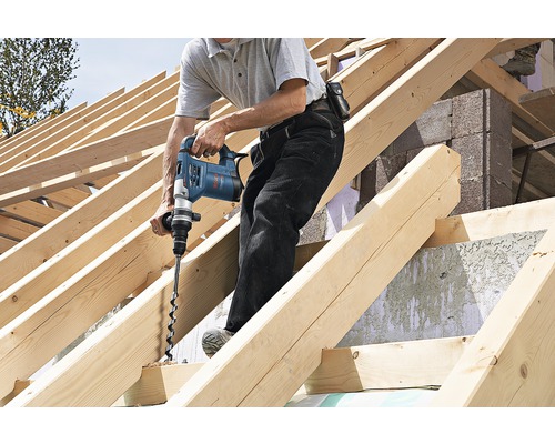 Mann bohrt mit einer Bohrmaschine ein Loch in einen Holzbalken auf einer Baustelle