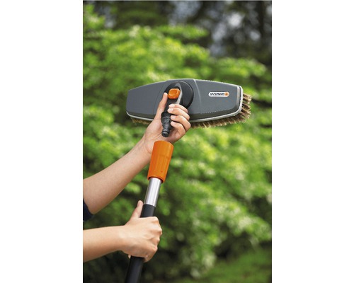 Une personne monte une brosse de lavage Gardena sur un manche télescopique pour des travaux de nettoyage dans le jardin.