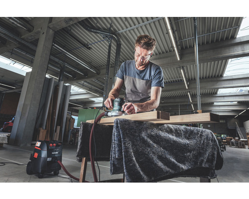 Un homme ponce une planche de bois avec une ponceuse excentrique dans un atelier, tandis qu''un aspirateur se trouve à côté.