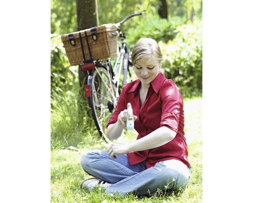 Une femme est assise à l''extérieur et se vaporise avec un insectifuge, à l''arrière-plan un vélo avec un panier.