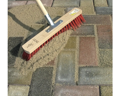 Eine Flächenbürste mit rotem Borstenbesatz kehrt Sand in die Fugen von Pflastersteinen.