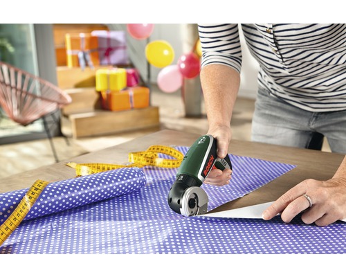 Scène avec une personne coupant du papier cadeau avec un cutter sans fil, ainsi qu''un mètre ruban et des emballages cadeaux