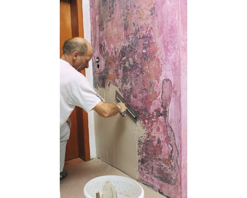 Handwerker glättet eine Wand mit Spachtelmasse vor einer renovierungsbedürftigen Wand.