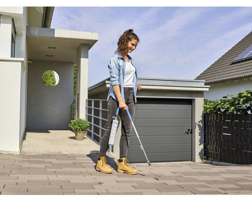 Une femme élimine les mauvaises herbes avec un brûleur à gaz sur une surface pavée devant une maison.