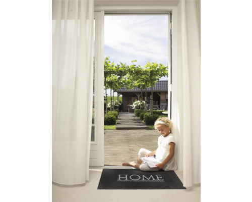 Une jeune fille assise sur un paillasson avec le mot ''Home'' devant une porte-fenêtre ouverte donnant sur un jardin, lit.