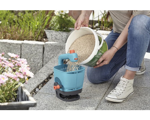 Eine Person befüllt einen Streuwagen mit Saatgut in einem Garten.