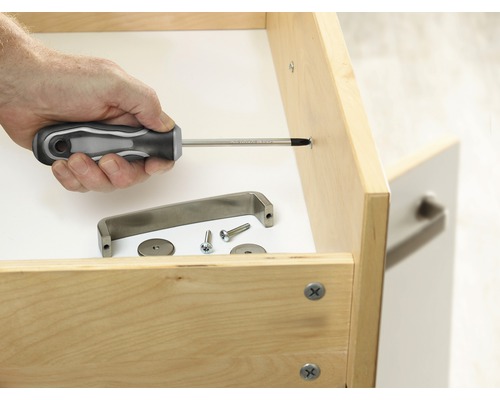 Une personne fixe une poignée de meuble à un tiroir avec un tournevis. Le matériel de montage est prêt.