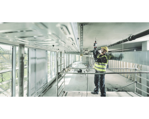 Ein Bauarbeiter auf einem Gerüst arbeitet mit einer Bohrmaschine an der Decke eines Rohbaus.