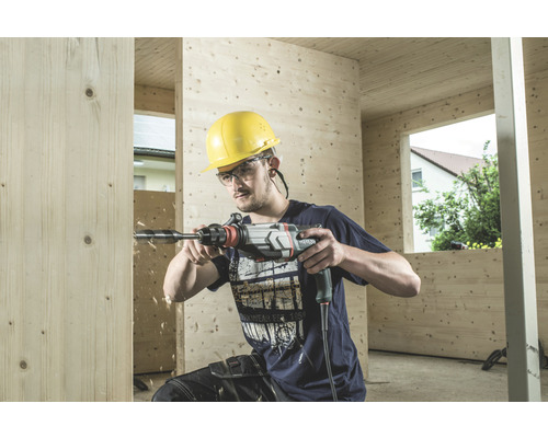 Mann bohrt mit einer Bohrmaschine ein Loch in eine Holzwand. Er trägt einen Schutzhelm und eine Schutzbrille.