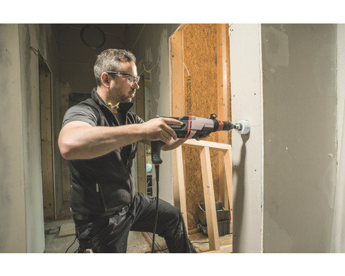 Ein Mann bohrt mit einer Bohrmaschine ein Loch in eine Wand. Die Szene zeigt eine Baustelle.