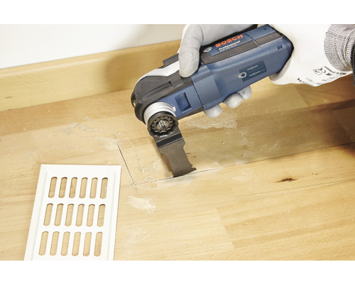 Une personne utilise un outil multifonction Bosch Professional pour découper une fente dans un plancher en bois à côté d''une grille de ventilation blanche.