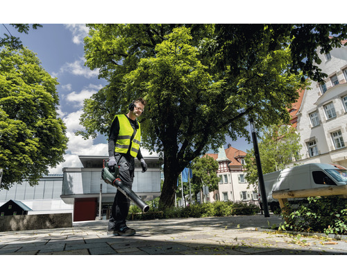 Ein Mann mit Sicherheitsweste und Kopfhörern benutzt einen Laubbläser im Freien.