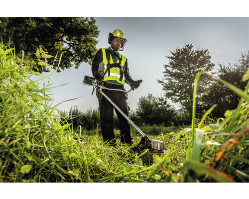 Ein Mann mäht hohes Gras mit einem Rasentrimmer und trägt dabei Schutzkleidung.