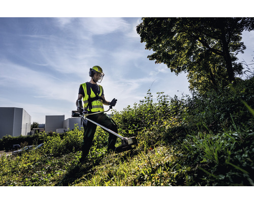 Mann arbeitet mit einem Rasentrimmer auf einer Wiese und trägt dabei Schutzkleidung.