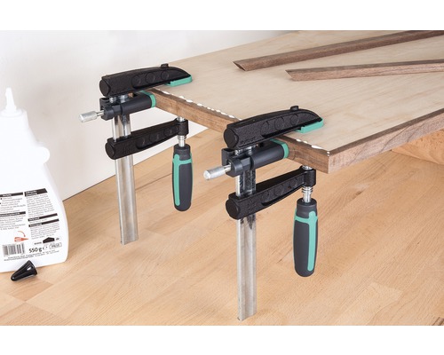 Auf dem Bild sind zwei Korpuszwingen zu sehen, die ein Holzbrett fixieren, das mit Holzleisten und Klebstoff versehen ist.