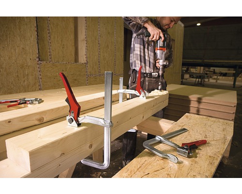 Mann bohrt mit einer Bohrmaschine durch ein Holzstück, das mit Schraubzwingen fixiert ist