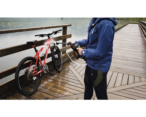 Une personne nettoie un vélo avec un nettoyeur haute pression portable sur un pont en bois près d''un point d''eau.