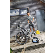 Ein Mann reinigt ein Fahrrad mit einem Hochdruckreiniger auf einer Holzterrasse vor einem Haus.