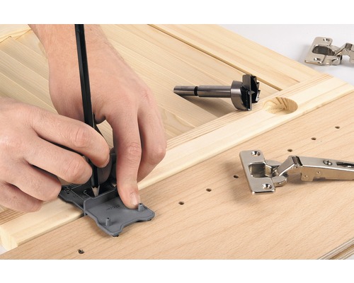 Une personne marque l''emplacement d''une charnière avec un gabarit et un crayon sur une planche de bois, à côté d''une mèche Forstner et d''une charnière.