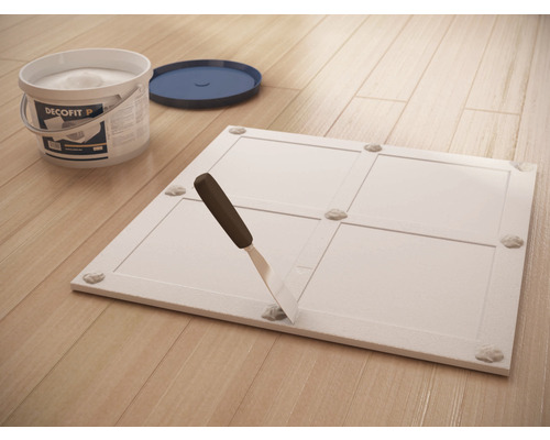 Dekorplatte mit Klebstoffpunkten, Spachtel und Eimer auf Holzboden