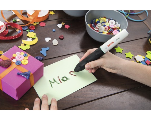 Person schreibt mit einem Klebestift auf eine Karte, umgeben von Bastelmaterialien und Geschenk
