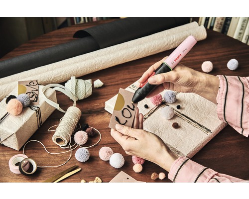 Bosch Klebepistole im Einsatz zum Beschriften eines Geschenkanhängers mit Geschenkpapier, Bändern und Bommeln auf einem Tisch