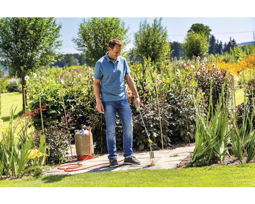 Ein Mann entfernt Unkraut mit einem Gasbrenner auf einer Steinterrasse in einem Garten.