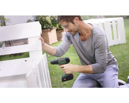 Homme portant des lunettes de sécurité utilisant une perceuse pour assembler une étagère en bois blanc dans le jardin. Logo Bosch.