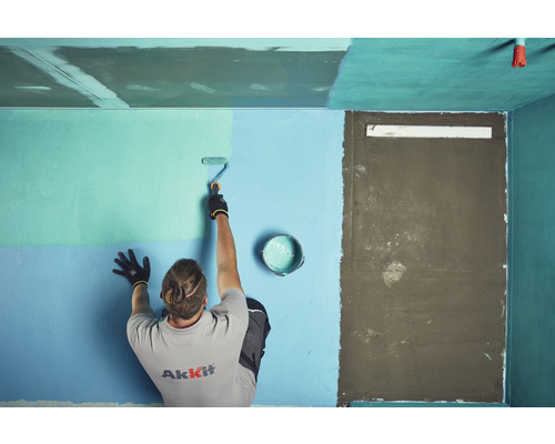 Un homme peint un mur avec un rouleau à peinture dans une pièce. Un pot de peinture est posé au sol.