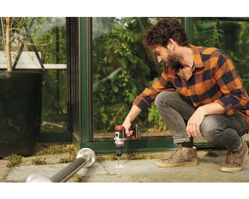 Un homme perce un trou dans le sol devant une serre avec une perceuse sans fil.