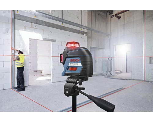 Logo Bosch. Chantier avec laser multilignes sur trépied, projetant des lignes laser rouges sur les murs et le sol. Un ouvrier portant un casque et un gilet de sécurité aligne un rail métallique.