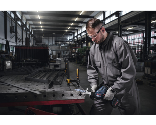Un homme portant des lunettes de protection et un casque antibruit travaille avec une scie sabre sans fil sur une pièce métallique dans un atelier.