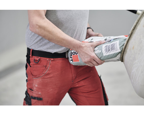 Mann mit roter Arbeitshose trägt einen Sack Baumaterial.