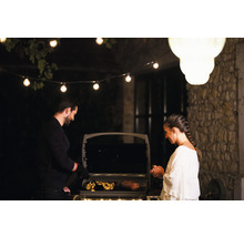 Un homme et une femme grillent de la viande et des pommes de terre sur un barbecue extérieur, éclairé par une guirlande lumineuse.