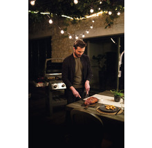 Homme coupant de la viande sur une terrasse avec un barbecue et une guirlande lumineuse