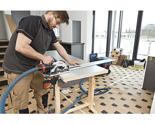 Un artisan coupe une pièce à usiner avec une scie circulaire à main sur un établi. Un tuyau d'aspiration est fixé à la scie.