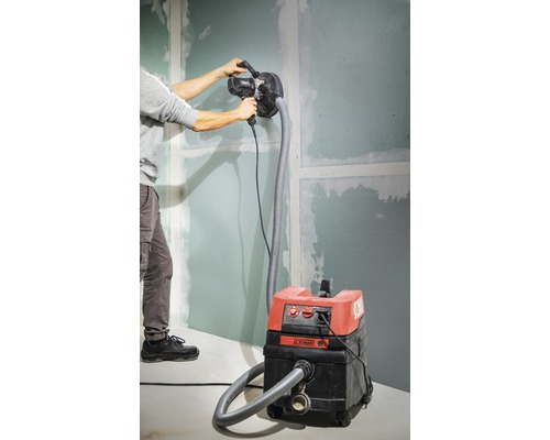 Un homme ponce un mur avec une ponceuse à plaques de plâtre raccordée à un aspirateur.