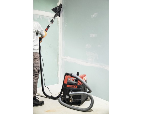 Eine Person schleift eine Wand mit einem Trockenbauschleifer und einem Staubsauger.