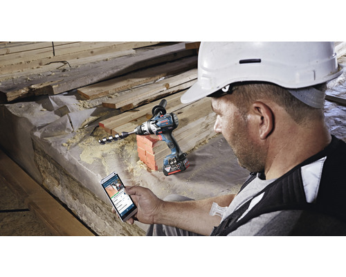 Handwerker mit Smartphone neben einem Akku-Bohrhammer auf einer Baustelle