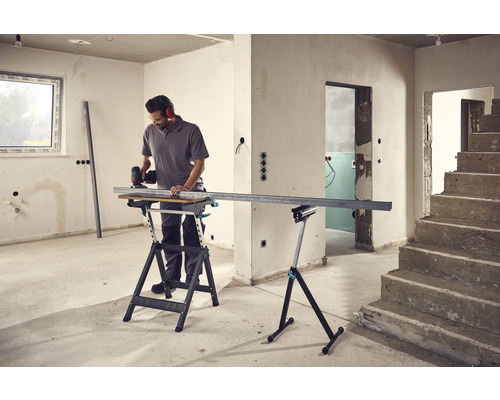 Un homme coupe un profilé métallique sur un établi dans une pièce non rénovée.