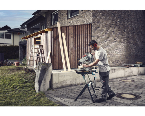 Un homme coupe du bois avec une scie à onglet dans le jardin devant une cabane en bois.