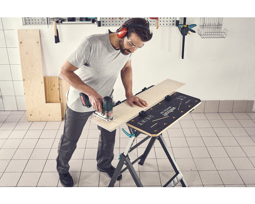 Un homme scie une planche de bois avec une scie sauteuse sur un établi. Il porte des lunettes de protection et un casque antibruit.