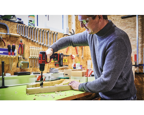Mann bohrt mit Akkubohrer ein Loch in ein Holzbrett in einer Werkstatt.