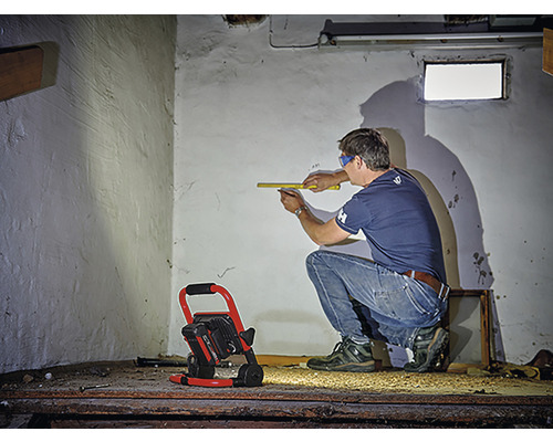 Un homme mesure un mur avec un mètre pliant, un projecteur de chantier éclaire la pièce