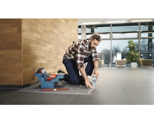 Un artisan agenouillé sur un sol carrelé travaille sur un profilé de transition. Une boîte à outils et un niveau à bulle sont à portée de main.