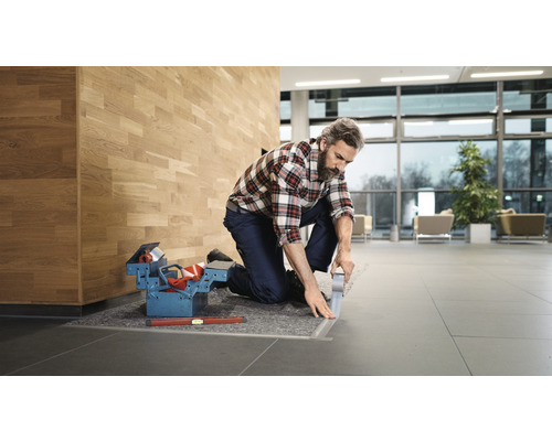 Un artisan agenouillé fixe un rail de sol avec du ruban adhésif, une boîte à outils et un niveau à bulle à côté.