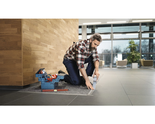Ein Handwerker kniet und verklebt eine Bodenschiene in einem Gebäude. Werkzeugkasten und Wasserwaage liegen bereit.