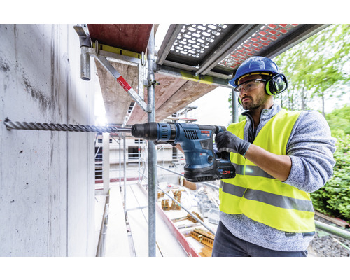 Un homme perce un trou dans un mur en béton sur un échafaudage avec un marteau perforateur.