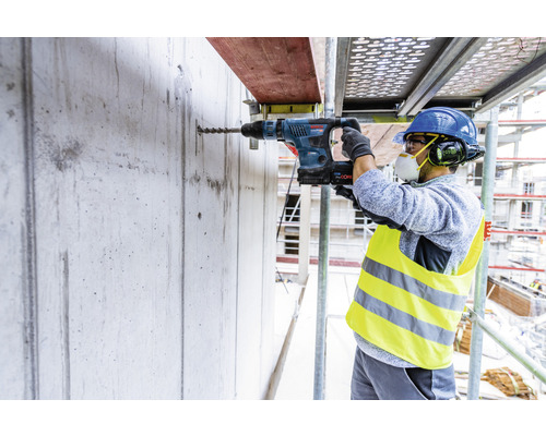 Un ouvrier perce un trou dans un mur en béton sur un échafaudage à l'aide d'un perforateur.