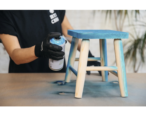 Une personne vaporise de la peinture bleue sur un petit tabouret en bois.